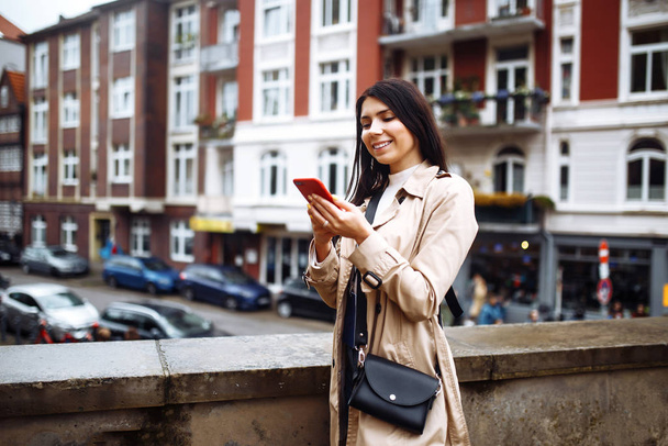 Mujer feliz con teléfono móvil caminando por la calle de Noruega. Hermosa chica elegante fotografía la arquitectura escandinava. Viajar, estilo de vida, aventura, concepto
. - Foto, imagen