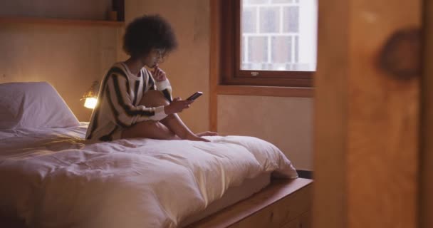 Side view of a young mixed race woman using a smartphone sitting on her bed at home - Video, Çekim