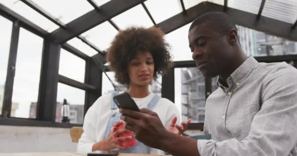  Mischlingsfrau mit Afrohaaren und ein afrikanisch-amerikanischer Mann sitzen an einem Tisch und trinken Kaffee, reden und lächeln in einem gläsernen überdachten Raum auf einem Dach mit städtischen Gebäuden im Hintergrund, schauen auf ein Smartphone, das der Mann in der Hand hält, Zeitlupe - Filmmaterial, Video