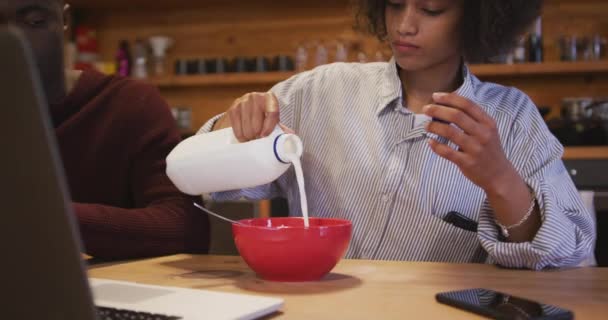 Front view of an African American man and a young mixed race woman with afro hair talking, eating breakfast cereal and using a laptop computer sitting at their kitchen table at home in the morning - Materiaali, video