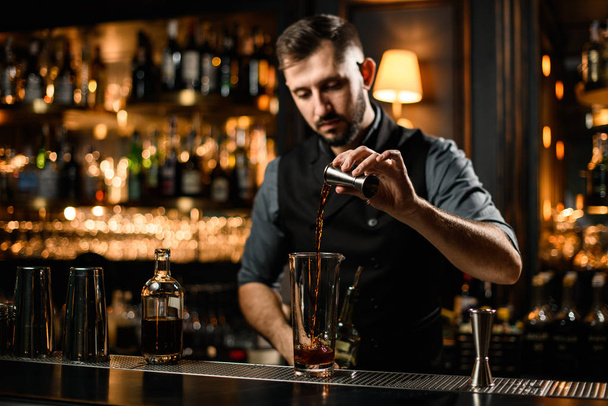 Professional bartender pouring a brown alcoholic drink from the jigger to a cocktail glass - Foto, Bild