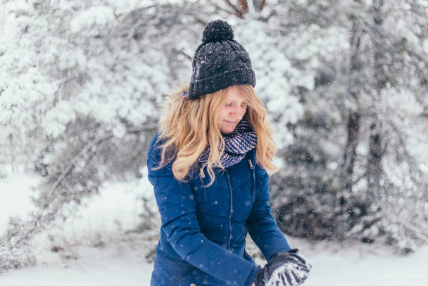 Close-up of happy cute attractive girl in woolen scarf enjoying winter moments - outdoor of long-haired blonde laughing lady in knitted hat sculpts a snowball in snowy day on blurred nature forest background - Φωτογραφία, εικόνα
