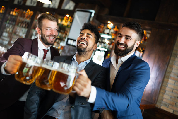 Handsome businessmen drinking beer, talking and smiling while resting at the pub - Fotó, kép
