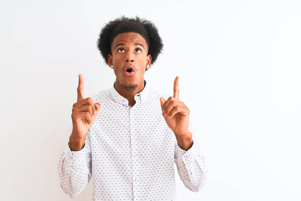 Jeune homme afro-américain portant une chemise élégante debout sur fond blanc isolé étonné et surpris regardant vers le haut et pointant avec les doigts et les bras levés
. - Photo, image