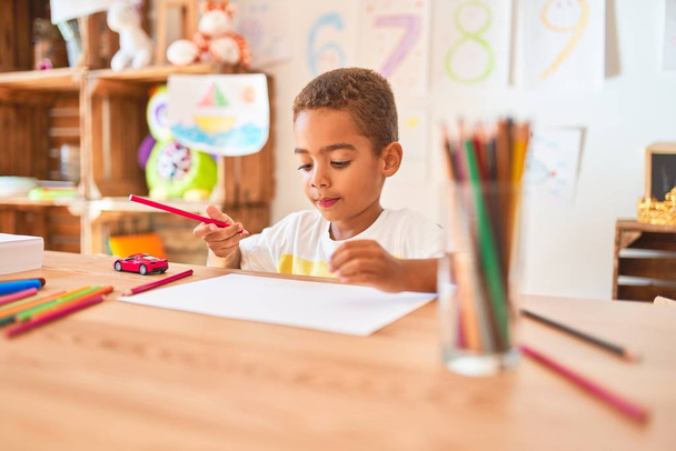 Beau bambin afro-américain assis à dessiner en utilisant du papier et des crayons sur le bureau à la maternelle
 - Photo, image