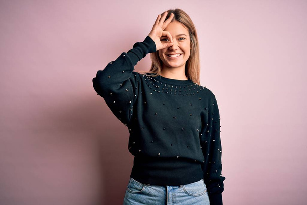 Young beautiful blonde woman wearing fashion sweater over pink isolated background doing ok gesture with hand smiling, eye looking through fingers with happy face. - Photo, image