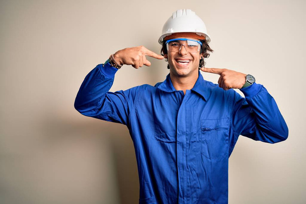Jonge constructeur man draagt uniform en veiligheidshelm over geïsoleerde witte achtergrond lachend vrolijk tonen en wijzen met vingers tanden en mond. Tandheelkundige gezondheidszorg. - Foto, afbeelding