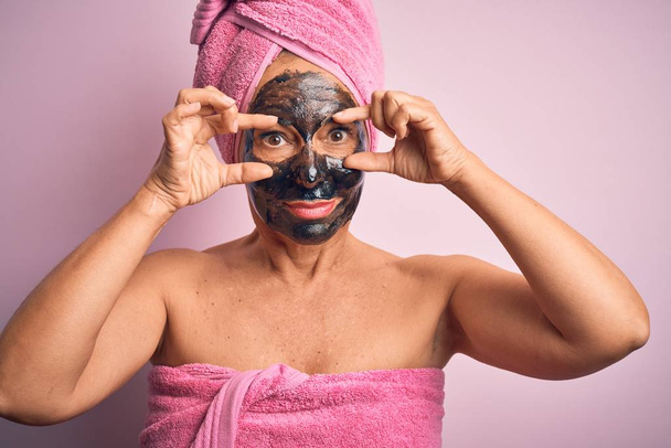Middelbare leeftijd brunette vrouw dragen schoonheid zwart gezicht masker over geïsoleerde roze achtergrond Proberen om ogen te openen met vingers, slaperig en moe voor 's morgens vermoeidheid - Foto, afbeelding