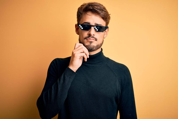Young man wearing thug life fanny sunglasses standing over isolated yellow background with hand on chin thinking about question, pensive expression. Smiling with thoughtful face. Doubt concept. - 写真・画像