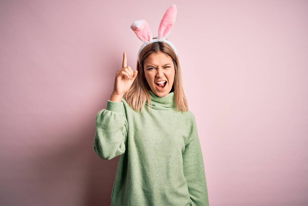 Молода красива жінка в великодніх вухах кролика стоїть над ізольованим рожевим тлом, вказуючи пальцем вгору з успішною ідеєю. Вийшов і щасливий. Номер один
. - Фото, зображення