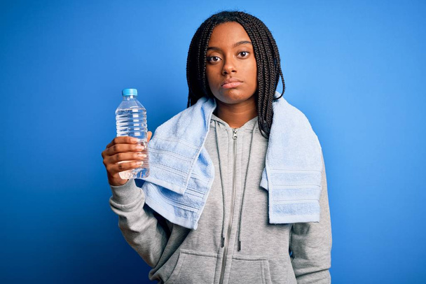 Joven afroamericana fitness mujer usando toalla y beber agua de botella de plástico con una expresión de confianza en la cara inteligente pensando en serio
 - Foto, Imagen