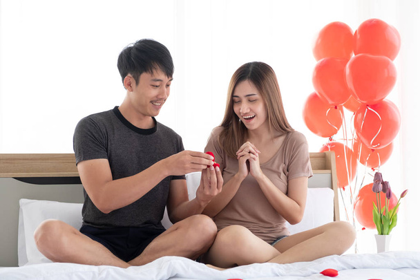 Le petit ami a surpris sa petite amie avec une bague contenue dans une boîte à bague rouge sur le lit dans la chambre décorée de ballons à cœur rouge pour la marier le jour de la Saint-Valentin
. - Photo, image