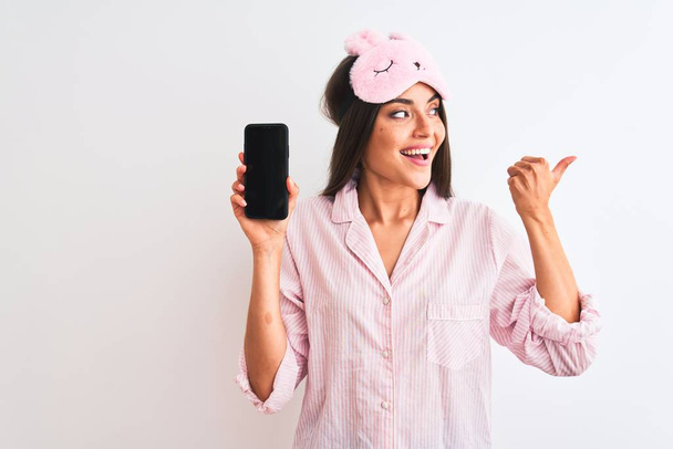 Young woman wearing sleep mask pajama holding smartphone over isolated white background pointing and showing with thumb up to the side with happy face smiling - Photo, Image