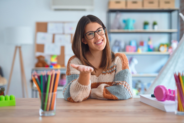 Młoda piękna nauczycielka nosząca sweter i okulary siedząca na biurku w przedszkolu uśmiechnięta radosna dłoń oferująca pomoc i akceptację. - Zdjęcie, obraz