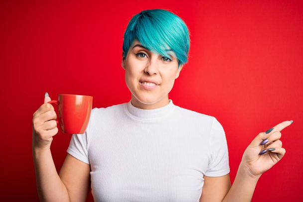 Молода жінка з синім модним волоссям п'є чашку кави над червоним ізольованим тлом дуже щаслива, вказуючи рукою і пальцем на бік
 - Фото, зображення
