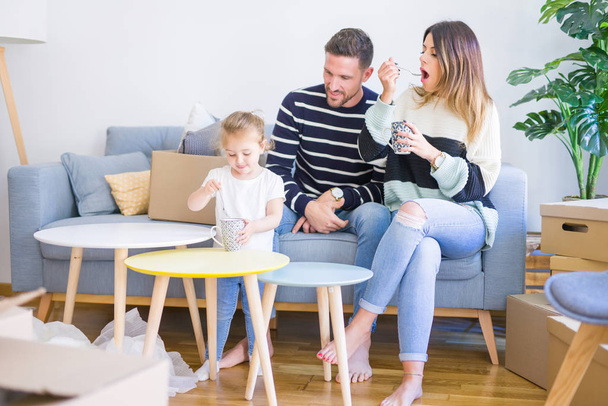 美しい家族、ソファに座ってコーヒーを飲む両親は、彼の子供が段ボール箱の周りに新しい家で遊んでいるのを見て - 写真・画像