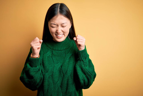 Fiatal gyönyörű ázsiai nő visel zöld téli pulóvert sárga elszigetelt háttér izgatott a siker karját, és csukott szemmel ünnepli győzelem mosolyogva. Nyertes koncepció. - Fotó, kép