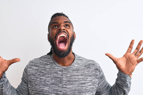 Afroamerikanischer Mann mit Zöpfen, grauem Pullover vor isoliertem weißen Hintergrund, verrückt und verrückt schreiend und schreiend mit aggressivem Ausdruck und erhobenen Armen. Frustrationskonzept. - Foto, Bild