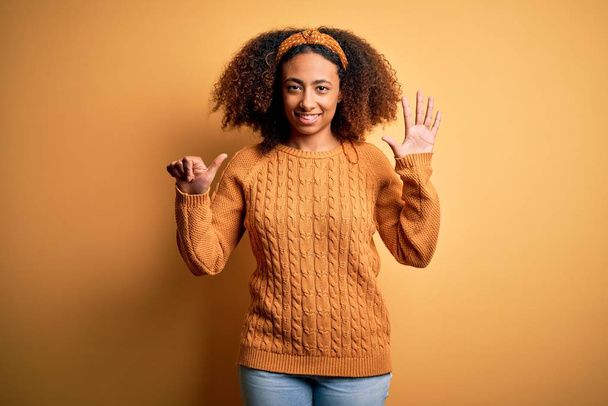 Fiatal afro-amerikai nő afro haj visel alkalmi pulóver sárga háttér mutatja, és rámutatva ujjak száma 6 miközben mosolygós magabiztos és boldog. - Fotó, kép