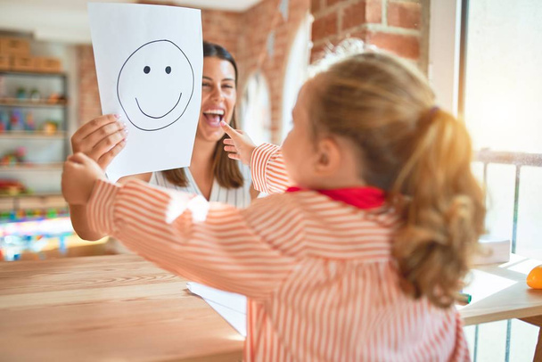 Piękny psycholog i blond dziewczynka maluch noszenie munduru szkoły siedzi na biurku robi terapię za pomocą emoji emocji w pokoju konsultacyjnym - Zdjęcie, obraz