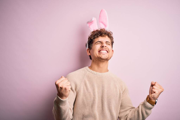 Young handsome blond man wearing easter rabbit ears over isolated pink background very happy and excited doing winner gesture with arms raised, smiling and screaming for success. Celebration concept. - Photo, Image