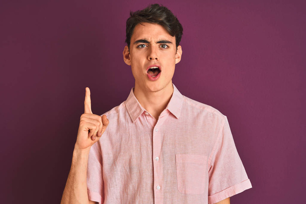 Teenager chlapec v růžové košili stojící nad fialové izolované pozadí ukazující prstem nahoru s úspěšným nápadem. Utekl a byl šťastný. Číslo jedna. - Fotografie, Obrázek