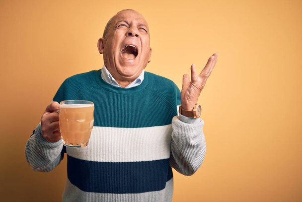 Senior handsome man drinking jar of beer standing over isolated yellow background crazy and mad shouting and yelling with aggressive expression and arms raised. Frustration concept. - Photo, Image