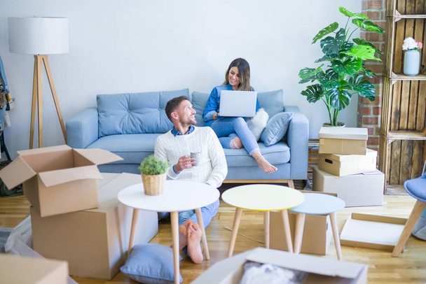 Young beautiful couple sitting on the sofa drinking cup of coffee using laptop at new home around cardboard boxes - Foto, Bild