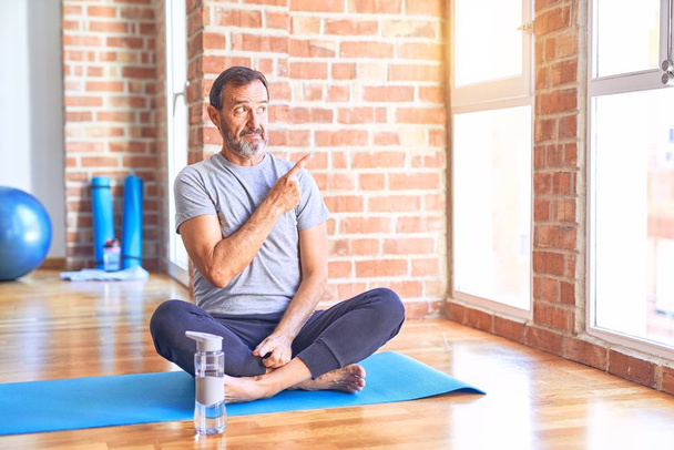 Middelbare leeftijd knappe sportman zittend op mat doen stretching yoga oefening in de sportschool Wijzen opzij bezorgd en nerveus met wijsvinger, bezorgd en verbaasd uitdrukking - Foto, afbeelding