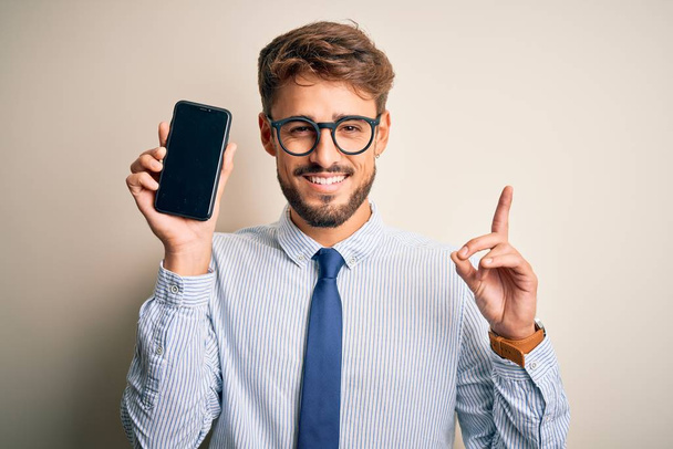 Giovane uomo d'affari che tiene lo smartphone in piedi su sfondo bianco isolato sorpreso da un'idea o domanda che punta il dito con la faccia felice, numero uno
 - Foto, immagini