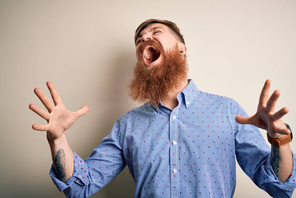 Handsome Irish redhead business man with beard standing over isolated background crazy and mad shouting and yelling with aggressive expression and arms raised. Frustration concept. - 写真・画像