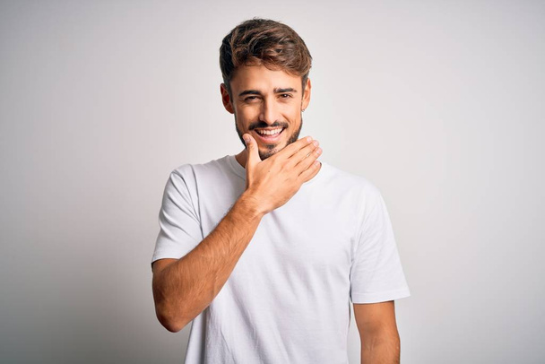 Young handsome man with beard wearing casual t-shirt standing over white background looking confident at the camera smiling with crossed arms and hand raised on chin. Thinking positive. - Foto, Imagen