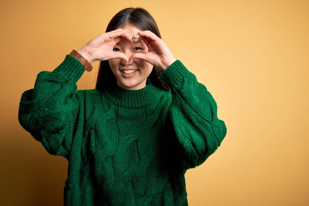 Young beautiful asian woman wearing green winter sweater over yellow isolated background Doing heart shape with hand and fingers smiling looking through sign - Foto, afbeelding