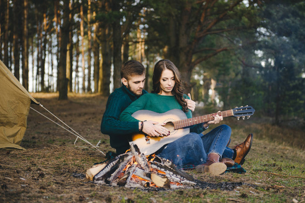 Young couple in love camping tourists sitting by a campfire against a tent in the woods playing guitar - Photo, image