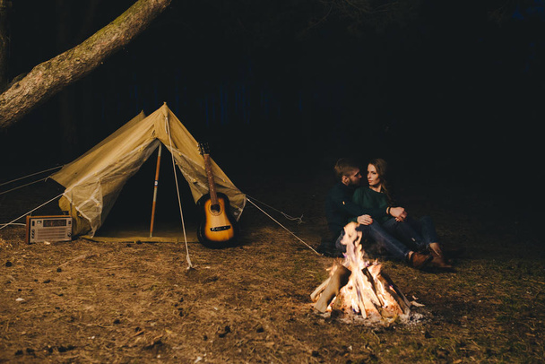 Young couple in love camping tourists sitting by a fire against a tent in the forest with a retro garland, photo with a lot of noise, selective focus - Photo, Image