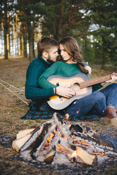 Young couple in love camping tourists sitting by a campfire against a tent in the woods playing guitar - Φωτογραφία, εικόνα