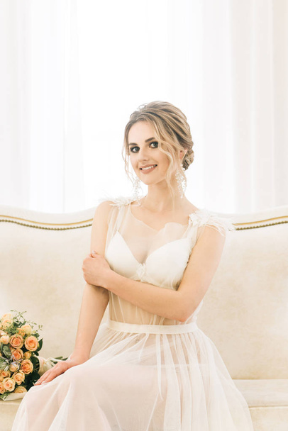 Πορτρέτο μιας όμορφης νεαρής νύφης σε ένα φωτεινό δωμάτιο σε μια ρομαντική ατμόσφαιρα. Νύφη σε νεγκλιζέ με γαμήλια ανθοδέσμη - Φωτογραφία, εικόνα
