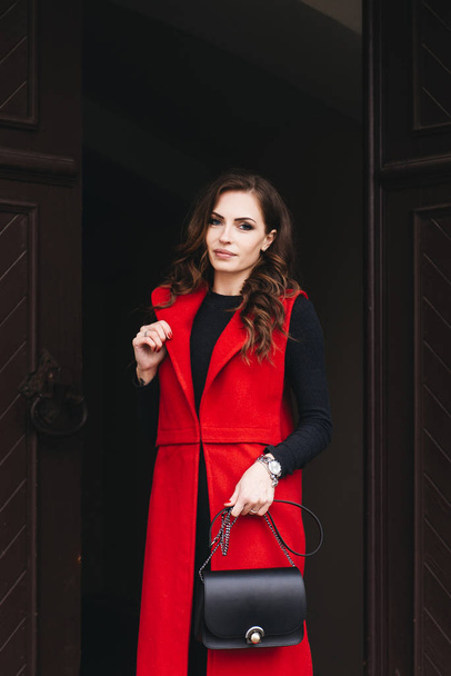 Outdoor portrait of a stylish young girl on the street of an old city in black clothes and a long red vest. A model poses on the street of a European city - Fotografie, Obrázek