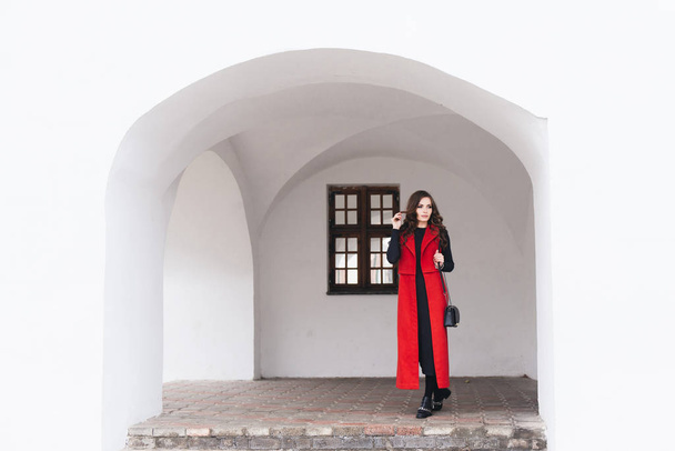 Outdoor portrait of a stylish young girl on the street of an old city in black clothes and a long red vest. A model poses on the street of a European city - 写真・画像