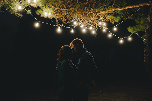 Giovane coppia innamorata campeggio turisti seduti vicino a un fuoco contro una tenda nella foresta con una ghirlanda retrò, foto con un sacco di rumore, messa a fuoco selettiva
 - Foto, immagini