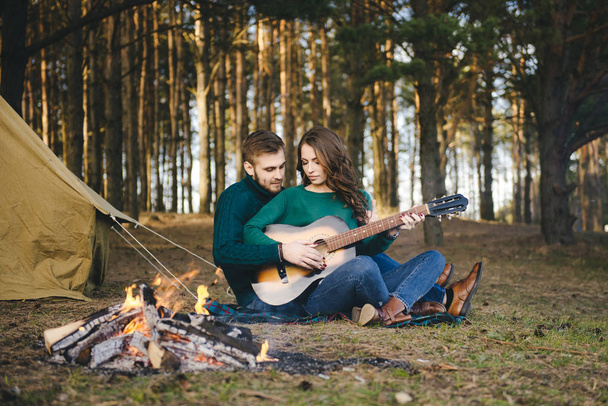 Young couple in love camping tourists sitting by a campfire against a tent in the woods playing guitar - Foto, afbeelding