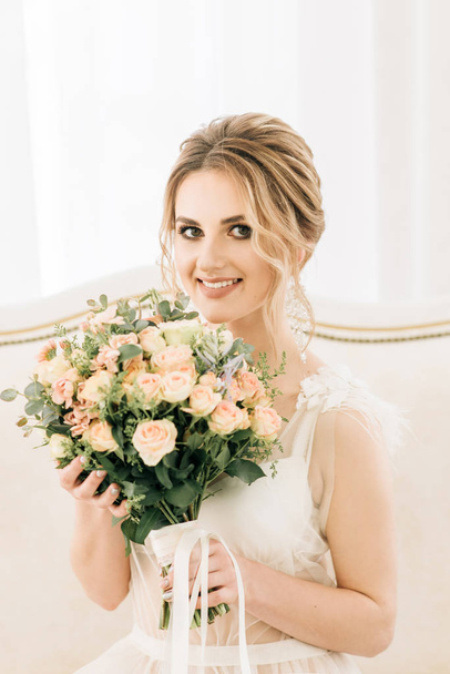 Portrait of a beautiful young bride in a bright room in a romantic atmosphere. Bride in a negligee with a wedding bouquet - Фото, изображение