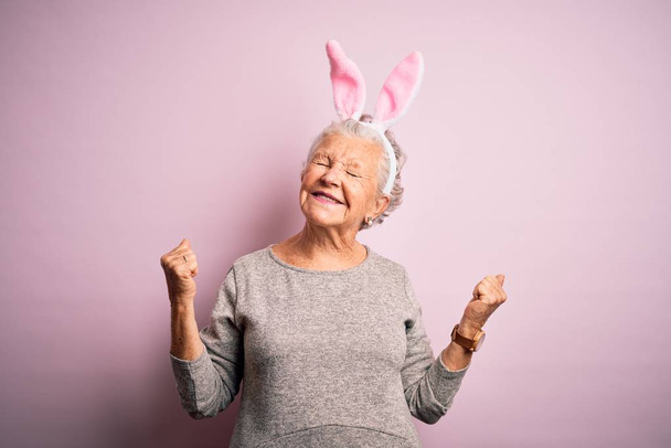 Starsza piękna kobieta nosząca królicze uszy stojąca nad odizolowanym różowym tle bardzo szczęśliwa i podekscytowana wykonując gest zwycięzcy z podniesionymi ramionami, uśmiechnięta i krzycząca o sukces. Koncepcja uroczystości. - Zdjęcie, obraz
