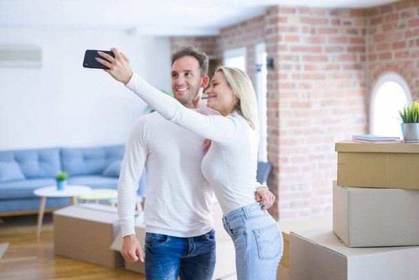 Joven hermosa pareja de pie utilizando el teléfono inteligente para tomar selfie en el nuevo hogar alrededor de cajas de cartón
 - Foto, Imagen