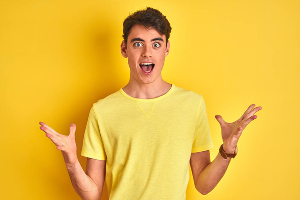 Adolescente niño vistiendo camiseta amarilla sobre fondo aislado celebrando loco y sorprendido por el éxito con los brazos levantados y los ojos abiertos gritando emocionado. Concepto ganador
 - Foto, imagen