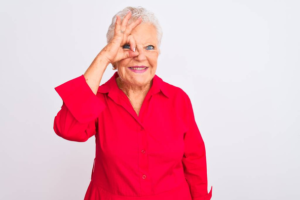 Femme âgée aux cheveux gris portant une chemise décontractée rouge debout sur un fond blanc isolé faisant un geste correct avec un sourire à la main, les yeux regardant à travers les doigts avec un visage heureux
. - Photo, image