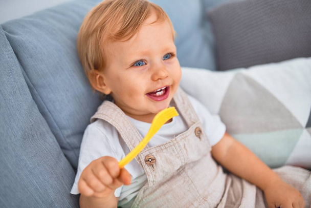 Adorable blonde toddler smiling happy sitting on the sofa playing with plastic meals toys at home - 写真・画像