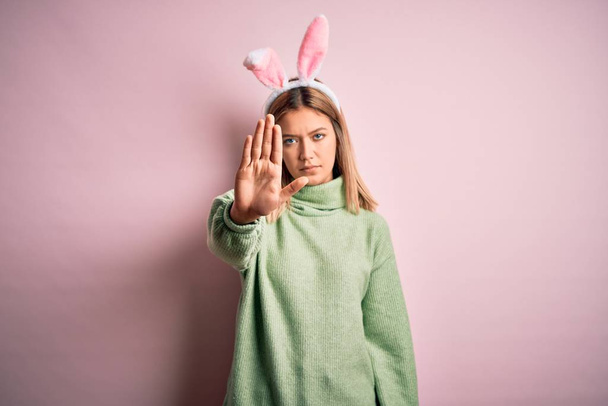 Young beautiful woman wearing easter rabbit ears standing over isolated pink background doing stop sing with palm of the hand. Warning expression with negative and serious gesture on the face. - Fotografie, Obrázek