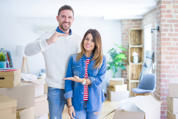 Young beautiful couple standing at new home around cardboard boxes gesturing with hands showing big and large size sign, measure symbol. Smiling looking at the camera. Measuring concept. - Photo, Image