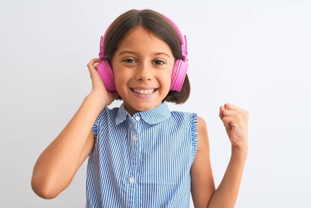 Όμορφο κορίτσι παιδί ακούγοντας μουσική χρησιμοποιώντας ακουστικά πάνω από απομονωμένο λευκό φόντο ουρλιάζοντας υπερήφανος και γιορτάζει τη νίκη και την επιτυχία πολύ ενθουσιασμένος, ζητωκραυγάζοντας συναίσθημα - Φωτογραφία, εικόνα